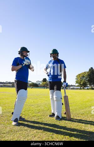 Diverse male teammates standing on cricket ground, wearing gloves, holding helmets and gripping bat. Athletes, teamspirit, sportsmanship, energetic, o Stock Photo