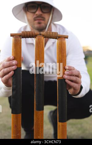 Man crouching and positioning wooden cricket stumps and bails wrapped in black tape on grassy field. Athletics, sportswear, minimalist, outdoor, teamw Stock Photo