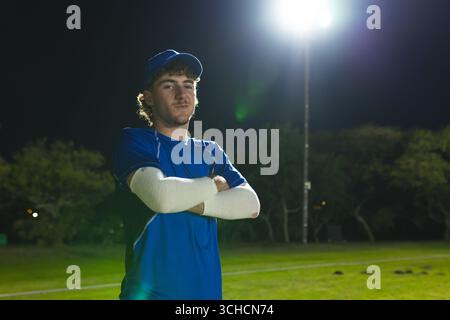 Male baseball player wearing blue cap and jersey standing on sports field under floodlight at night. Athlete, sports, recreation, outdoor, nighttime, Stock Photo