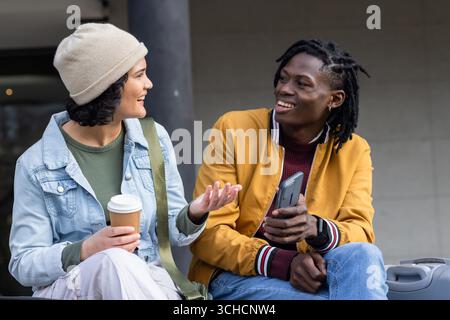 Diverse friends chatting outside building with coffee cup and checking phone by suitcase and bag. Casual, urban, modern, lifestyle, conversation, comp Stock Photo