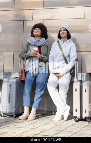 Diverse female friends standing on sidewalk holding coffee cup and smartphone next to suitcases. Casual, travel, companionship, urban, lifestyle, prep Stock Photo