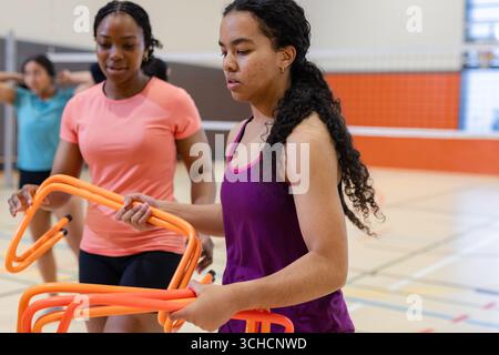 Diverse female teammates arranging orange agility hurdles along painted court lines in gym hall. Athletics, teamwork, training, fitness, collaboration Stock Photo