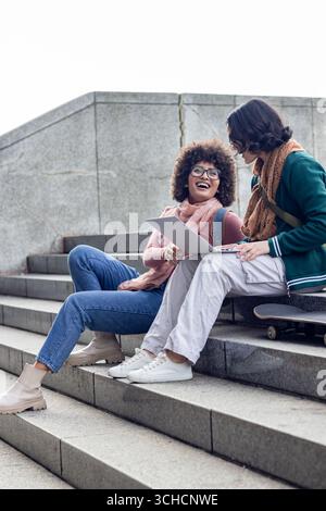 Female friends sitting on steps in urban plaza pointing at laptop holding tablet beside skateboard. Casual, friendship, collaboration, urban, youth, t Stock Photo