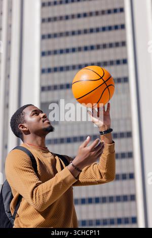 African American man standing outdoors, spinning basketball while wearing mustard sweater. Urban, professional, modern, vibrant, lifestyle, athletic, Stock Photo