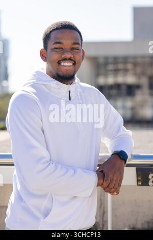 African American man leaning against terrace railing smiling sporting smartwatch. Urban, lifestyle, contemporary, technology, fashion, confidence, out Stock Photo