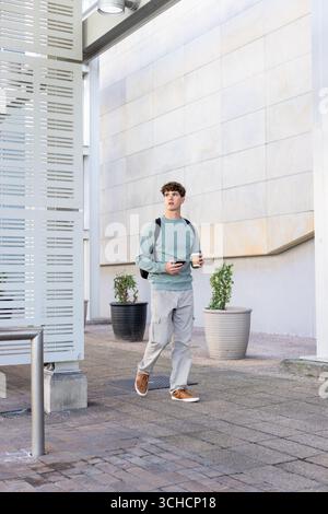 Male walking along paved plaza beside modern building carrying coffee cup smartphone and backpack. Urban, modern, contemporary, minimalism, profession Stock Photo