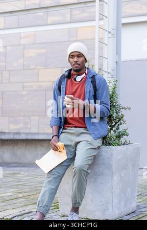 African American man sitting on edge of planter in courtyard with headphones holding coffee cup. Urban, casual, modern, lifestyle, youth, relaxation, Stock Photo