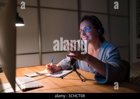 Smiling middle-aged woman in headphones chatting on video call using microphone with pop filter and desktop computer, internet communication technolog Stock Photo
