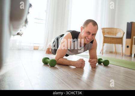 Home workout in living room smiling cheerful young muscular man doing plank exercise with dumbbells nearby promoting healthy lifestyle fitness motivat Stock Photo