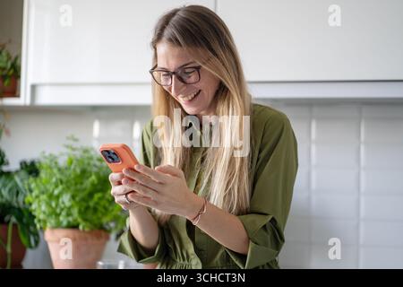 Positive woman typing messages or video calling on smartphone standing in home kitchen with smile, Stock Photo