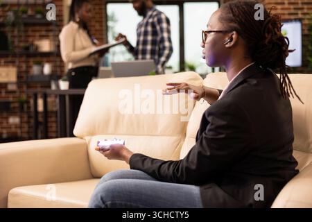 Businesswoman using futuristic hologram device to present data in modern workspace. Black female manager, wearing wireless earphones and seated on sofa, communicates with holographic interface Stock Photo
