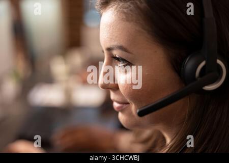 Extreme closeup of caucasian woman employee with headset providing online support, displaying professional expertise. Smiling female telemarketer on call, virtually assisting customer with an inquiry. Stock Photo