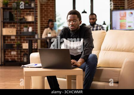 Focused black male manager sitting on couch and using laptop in brick wall office. African american businessman replying to emails and checking daily planner for productive day in modern workspace. Stock Photo