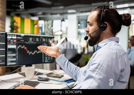 Professional consultant in office talking with clients, explaining stock market trends and investment options through headset microphone. Man answers customer questions and gives financial advice Stock Photo