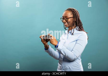 Cheerful black female model plays interactive mobile games on phone, fully engaged in the experience while standing in studio. Portrait of woman enjoying gaming apps on cellphone. Stock Photo