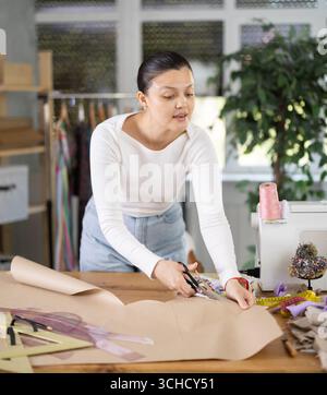 Smiling young woman seamstress cutting pieces of paper with scissors according to drawn template for pattern during dress design at workplace in Stock Photo