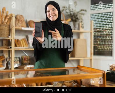 Female bakery owner in hijab promoting mobile ordering app Stock Photo