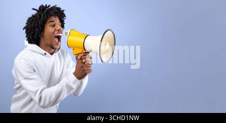 Emotional black teen guy shouting into megaphone, announcing breaking news over violet background Stock Photo