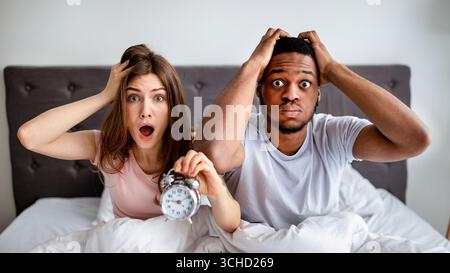 Shocked multiracial couple with alarm clock being late to work, sitting on bed, grabbing their heads in terror Stock Photo