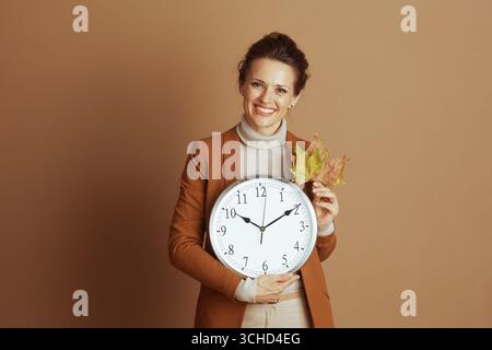 A cheerful businesswoman in a stylish blazer smiles at the camera, holding a large wall clock and autumn leaves, conveying punctuality, seasonal trans Stock Photo
