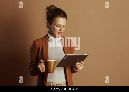 A focused businesswoman in a brown blazer enjoys a coffee break while using a digital tablet. Her calm, engaged demeanor captures the essence of remot Stock Photo