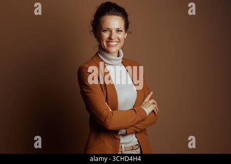 A confident businesswoman with a warm smile and arms crossed, wearing a brown blazer and beige turtleneck, looks directly at the camera against a soli Stock Photo