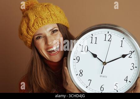 A cheerful woman in an orange turtleneck and yellow beret smiles broadly, holding a large wall clock. She embodies a happy, energetic interaction with Stock Photo