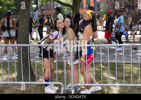 Three young women pose in front of a metal fence, two of them embracing. They are casually dressed, with one wearing a Puerto Rican flag-themed shirt Stock Photo