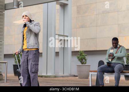 Two men standing outside building holding envelope and pulling suitcase, checking watch and phone Stock Photo