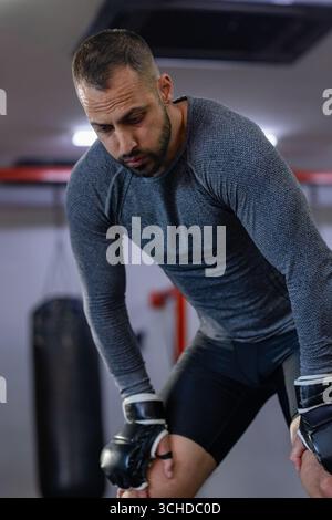 Man leaning with hands on thighs, catching breath on padded floor under punching bag Stock Photo