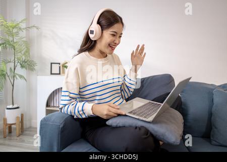 A young woman sitting on a sofa with headphones, smiling and gesturing happily while using a laptop. She looks excited and engaged, enjoying a joyful Stock Photo
