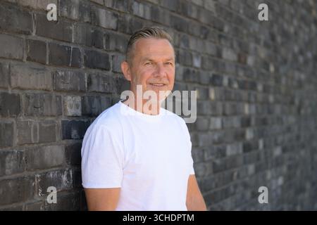 Portrait of a smiling senior man leaning casually against a brick wall. Stock Photo