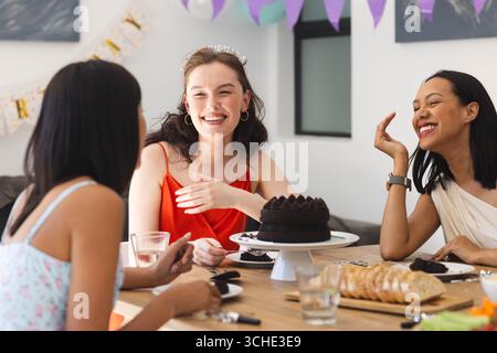 Diverse female friends celebrating at home, enjoying cake and laughter around festive table Stock Photo