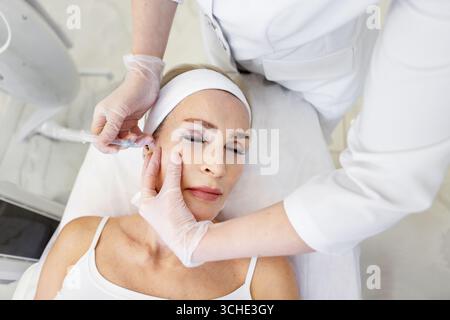 Senior woman receiving aesthetic facial injection by professional cosmetologist in white coat and gloves during anti-aging treatment procedure in mode Stock Photo