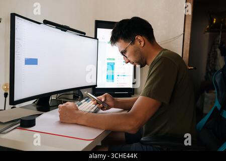 Programmer working from home office checking calendar on phone and scheduling meeting while sitting at desk in front of two computer screens. Stock Photo