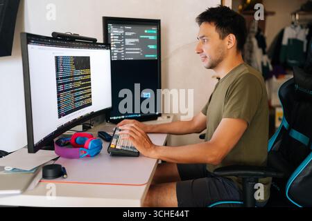 Focused software developer writing code at home office desk, surrounded by dual monitors displaying lines of code and communication platforms in Stock Photo
