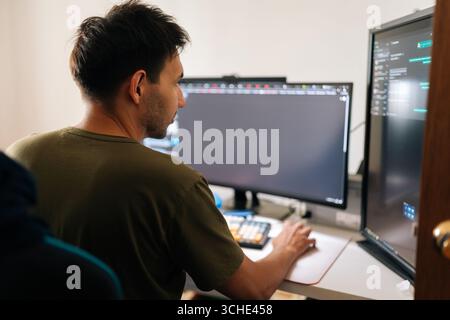 Close-up rear view of programmer male working from home office using multiple monitors setup and typing on mechanical keyboard looking to screen Stock Photo