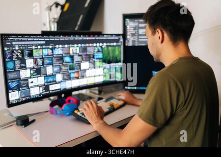 Back view of software developer male writing code at home office desk, surrounded by dual monitors displaying lines of code and communication Stock Photo