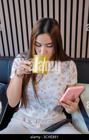 Young woman enjoys morning coffee while using smartphone on comfortable sofa, remote control nearby Stock Photo