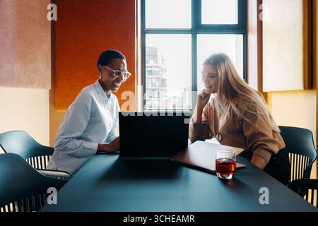Two young professional women in conversation while sitting in a bright office setting. Stock Photo