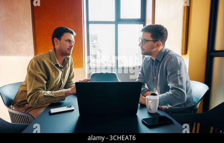 Two young adult men engage in a focused business discussion in a bright, professional office atmosphere. A laptop and smartphone on the table suggest Stock Photo