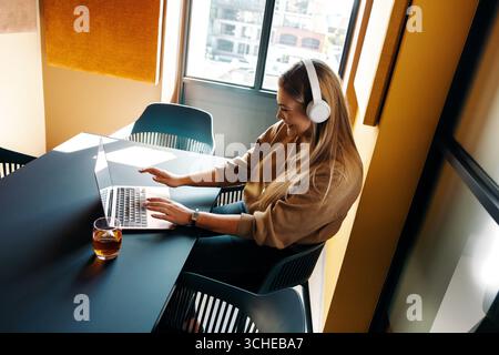 Smiling woman in her 20s with headphones sitting at a table using a laptop. Stock Photo