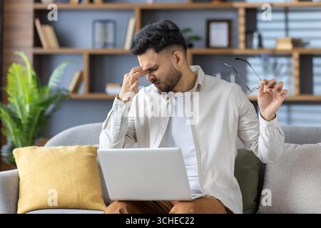 Man sitting on couch with laptop experiencing discomfort and eye strain, holding glasses and massaging forehead. Concept of remote work challenges, st Stock Photo