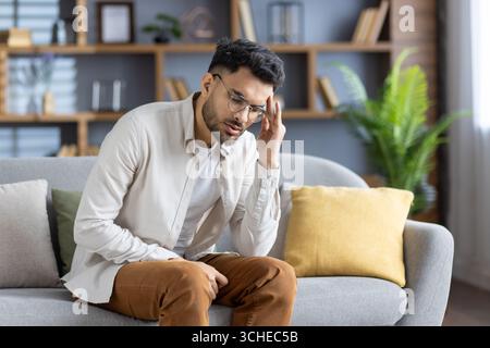 Young man with glasses experiencing headache sitting on sofa, displaying stress and fatigue. Casual setting at home, holding head in discomfort, weari Stock Photo