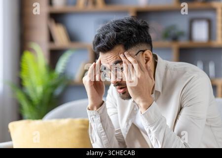 Man experiencing stress, sitting on couch holding head, coping with difficult emotions in home office. Overwhelmed, wearing glasses, casual clothes, c Stock Photo