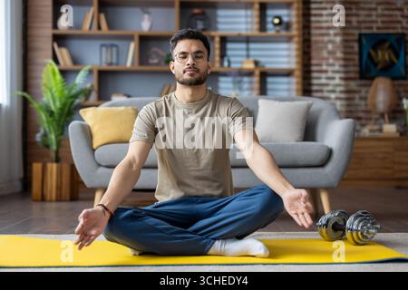 Man sitting cross-legged on yoga mat meditating in living room. Practicing mindfulness and relaxation. Comfortable home setting with dumbbells indicat Stock Photo