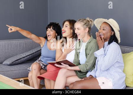 Diverse group of diverse female friends on couch, laughing and pointing at something, at home Stock Photo