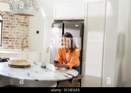 At home, woman using smartphone in modern kitchen, sitting at marble counter Stock Photo