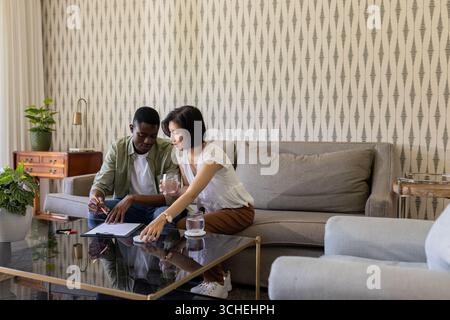 Reviewing paperwork Diverse couple sitting on sofa in living room, with glass-top table and pen Stock Photo