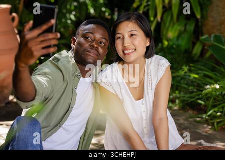 Taking selfie Diverse couple sitting on stone patio in lush garden, with smartphone Stock Photo